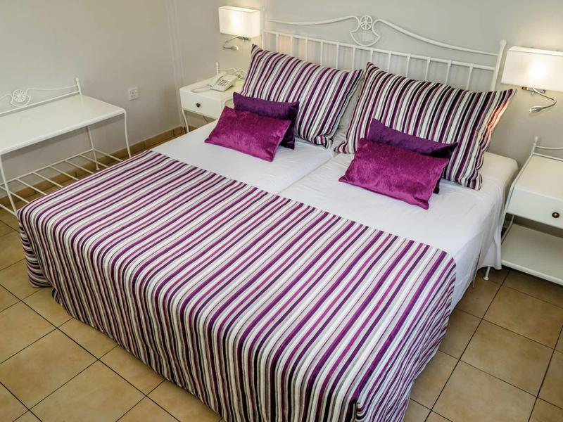 Doppelbett mit gestreifter Bettwäsche in Weiß, Lila und Pink, daneben weiße Nachttische mit Lampen.