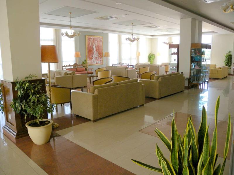 Großer, heller Aufenthaltsraum mit beigen Sofas, Pflanzen und viel Tageslicht durch große Fenster.