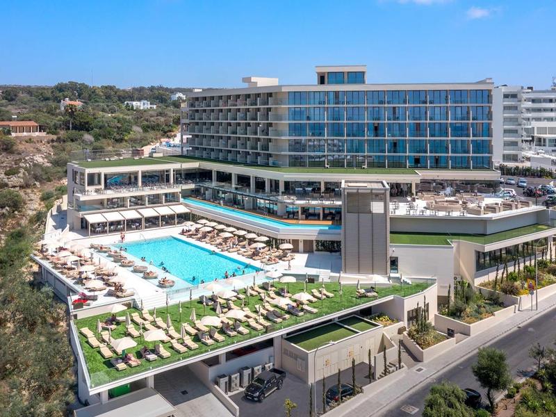 Modernes Hotel mit großem rechteckigem Pool, Sonnenliegen und Panoramafenstern in sonniger Landschaft.