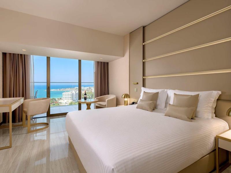 Helles Hotelzimmer mit großem Bett, Balkontür mit Meerblick, Schreibtisch und zwei Stühlen.