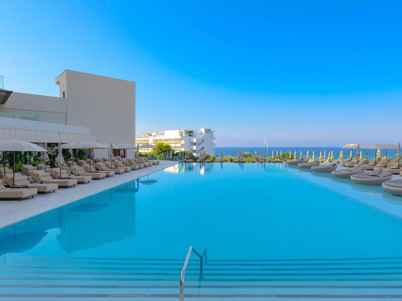 Großer blauer Pool mit vielen Liegestühlen und klar blauem Himmel am Meer im Hintergrund.