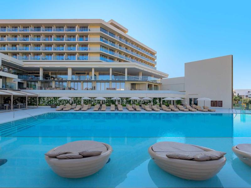 Moderner Hotelkomplex mit großem Pool, Sonnenliegen und klar blauem Himmel im Hintergrund.
