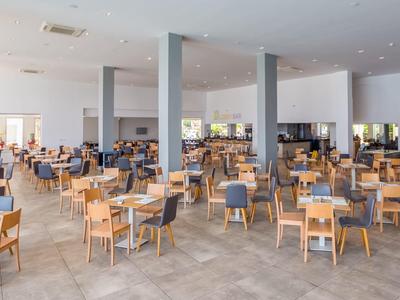 Großer, heller Speisesaal mit vielen Holzstühlen und grauen Polstersitzen, heller Boden und weiße Wände.