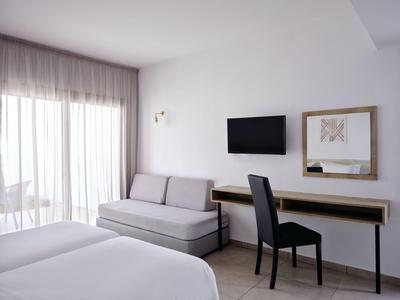 Helles Zimmer mit zwei Betten, grauem Sofa, schwarzem Stuhl, Wandfernseher und großem Fenster.