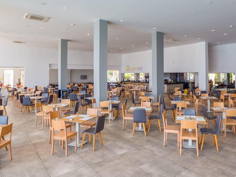 Großer, heller Speisesaal mit vielen Holzstühlen und grauen Polstersitzen, heller Boden und weiße Wände.