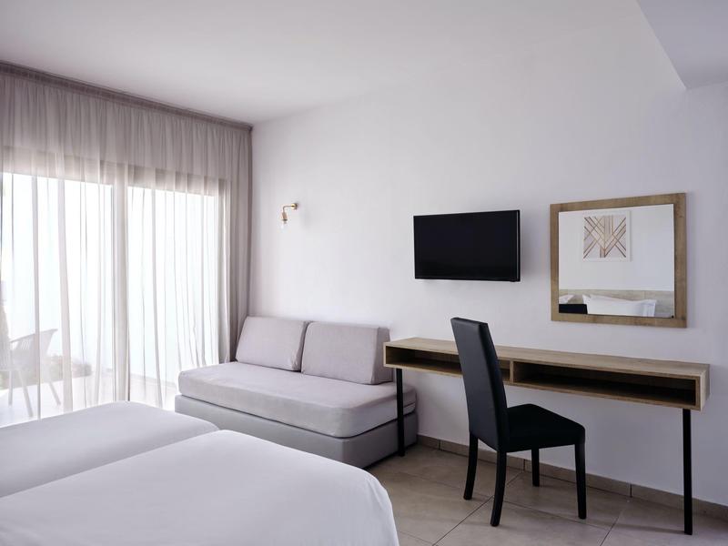 Helles Zimmer mit zwei Betten, grauem Sofa, schwarzem Stuhl, Wandfernseher und großem Fenster.