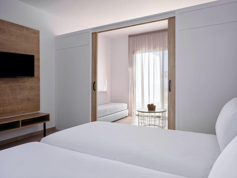 Modernes Schlafzimmer mit weißem Bett und Schiebetür zum hellen Wohnzimmer mit Sofa.