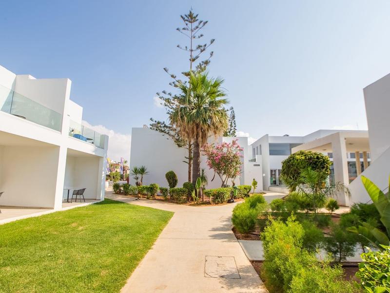 Wohnanlage mit weißen Gebäuden, gepflegtem Garten, Palmen und Gehweg unter blauem Himmel.