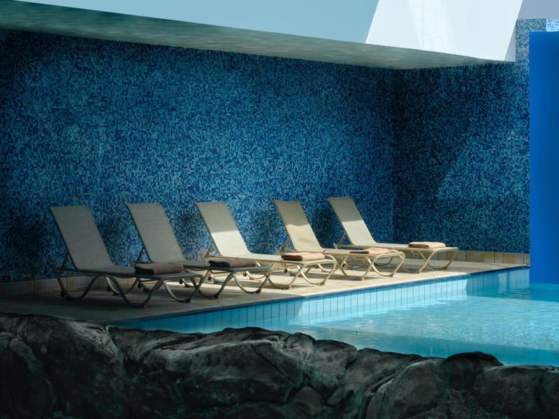 Innenpoolbereich mit blauer Mosaikwand und sechs Liegestühlen in Reihenanordnung entlang des Beckens.