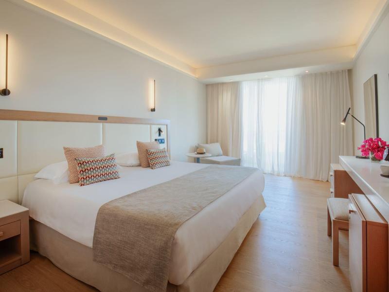 Chambre d'hôtel lumineuse avec lit double, bureau, chaise et grande fenêtre avec rideaux.