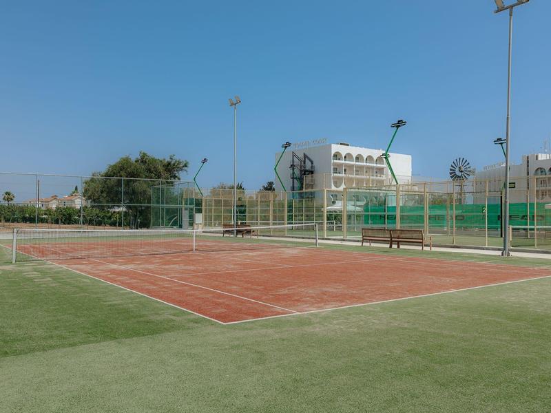 Court de tennis extérieur avec surface rouge et bord vert sous un ciel bleu clair.