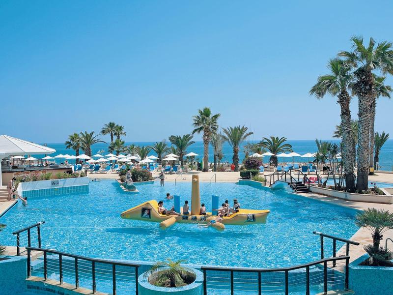 Piscine d'hôtel avec chaises longues et palmiers, vue sur la mer par temps ensoleillé.