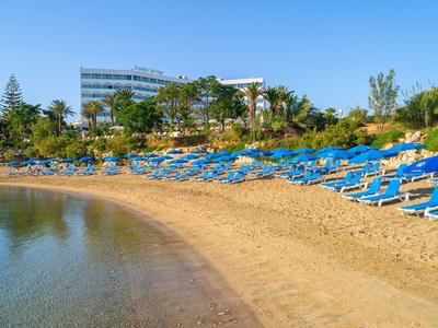 Plage de sable avec des chaises longues bleues devant un hôtel et une végétation verte.