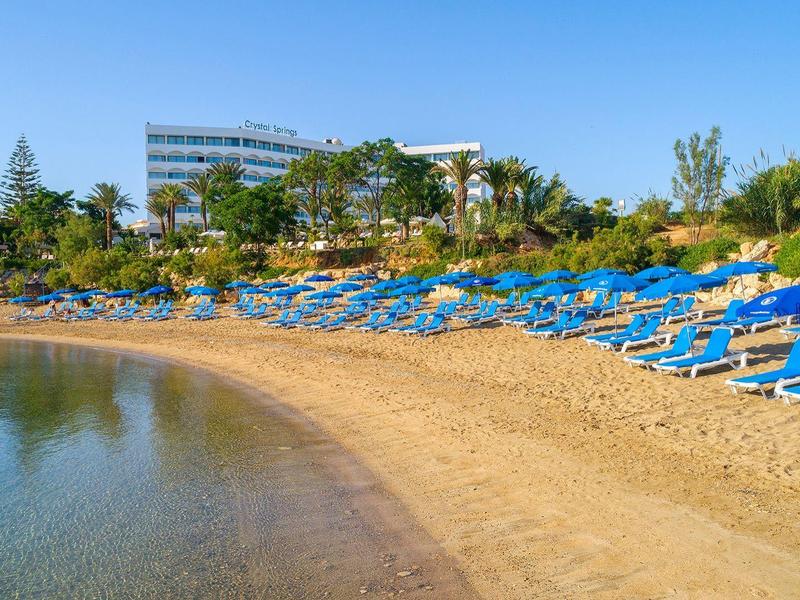 Plage de sable avec des chaises longues bleues devant un hôtel et une végétation verte.