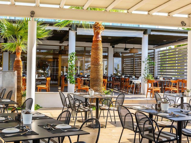 Espace extérieur moderne d’un restaurant avec tables, chaises et palmiers sous une pergola.