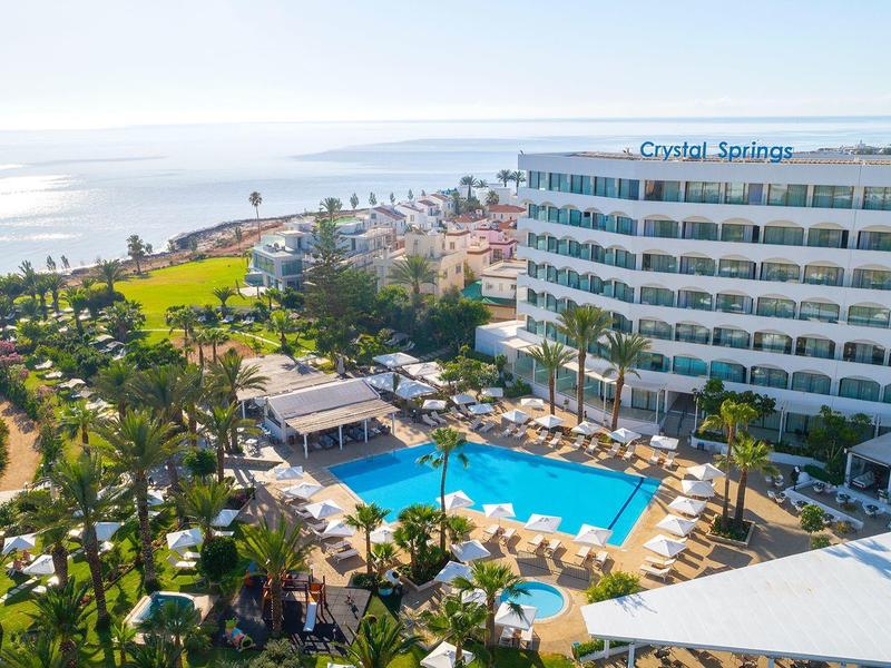 Grand hôtel avec piscine extérieure, chaises longues et vue sur la mer sous un ciel clair.