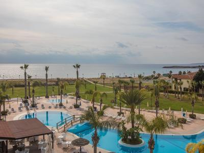 Blick auf einen Hotelpool mit Palmen, Liegestühlen und Blick auf das Meer bei bewölktem Himmel.