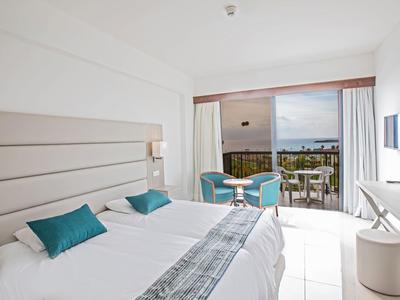 Helles Hotelzimmer mit Doppelbett, blauen Kissen und Balkon mit Meerblick.