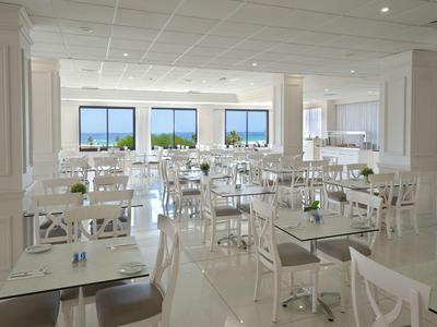 Heller, moderner Speisesaal mit weißen Tischen und Stühlen, großen Fenstern mit Meerblick.