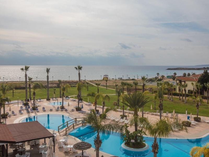 Blick auf einen Hotelpool mit Palmen, Liegestühlen und Blick auf das Meer bei bewölktem Himmel.