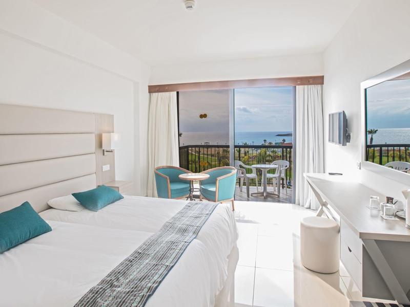 Helles Hotelzimmer mit Doppelbett, kleinem Tisch, Stühlen und Balkon mit Meerblick.