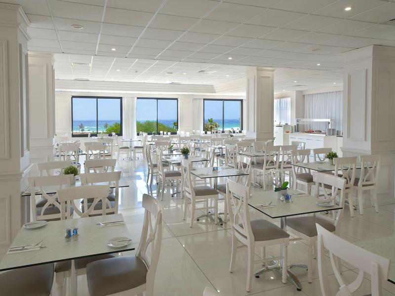 Heller, moderner Speisesaal mit weißen Tischen und Stühlen, großen Fenstern mit Meerblick.