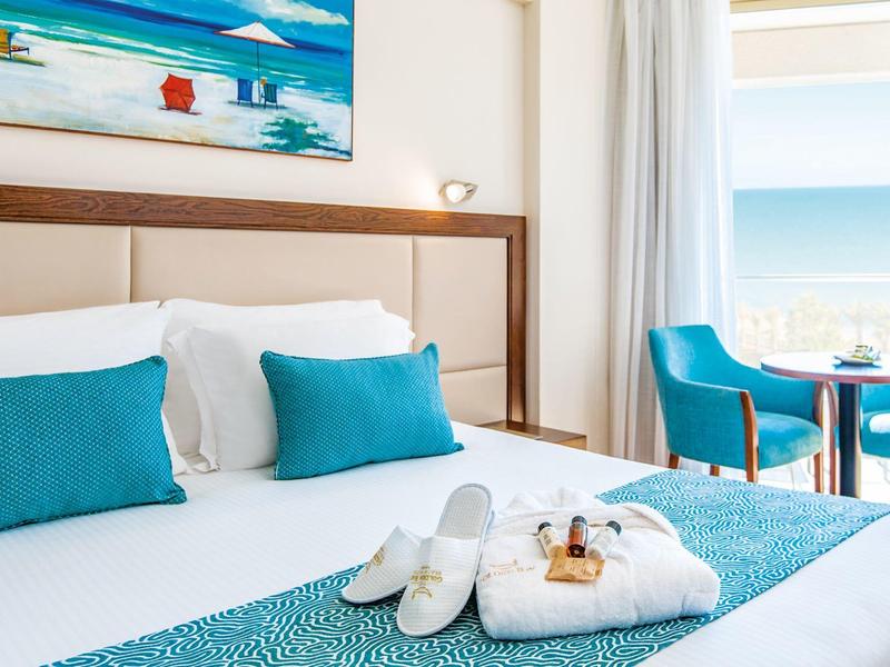 Helles Hotelzimmer mit Meerblick, weißem Bett, türkisfarbenen Kissen und Stühlen, Handtüchern und Slippern.