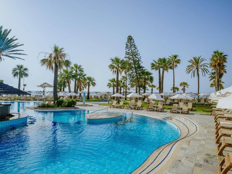Großer Pool mit blauem Wasser, Palmen drumherum und Liegestühlen an sonnigem Himmel.