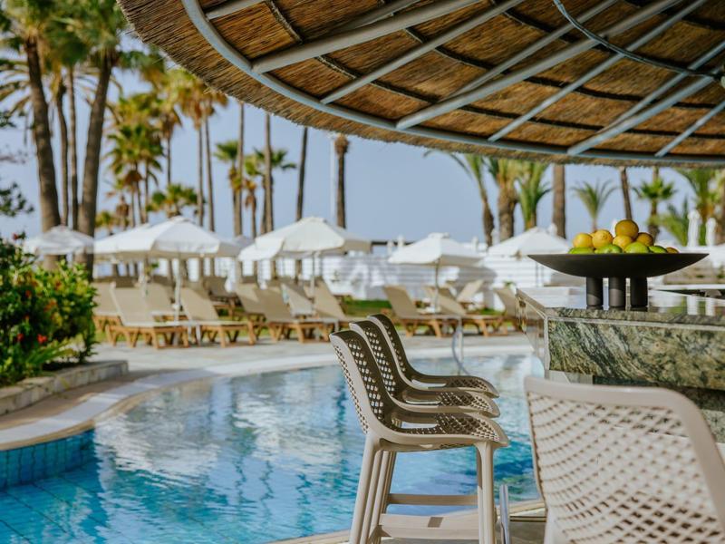Poolbar mit weißen Stühlen und Sonnenschirmen, Palmen und blauem Wasser im Hintergrund.