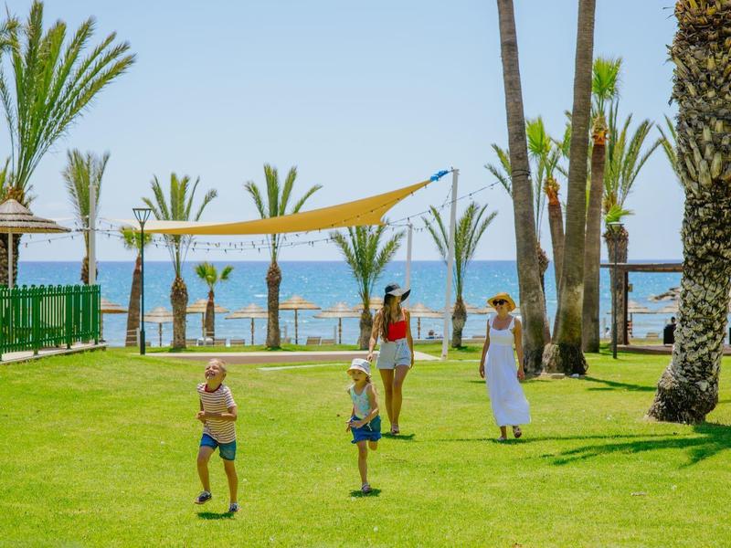 Grüne Wiese mit Palmen, Familie auf Spaziergang, blauer Himmel und Meer im Hintergrund.