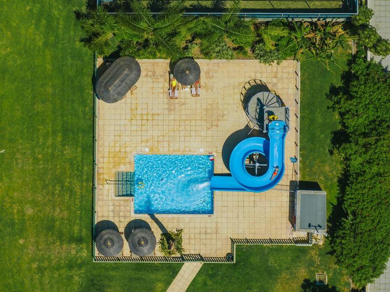 Vogelperspektive eines Pools mit blauer Rutsche und umliegender Terrasse aus hellen Fliesen.