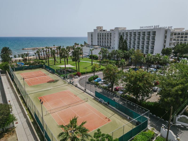 Mehrere Tennisplätze mit rotem Sand, umgeben von grünen Büschen, Palmen und einem großen weißen Gebäude am Meer.