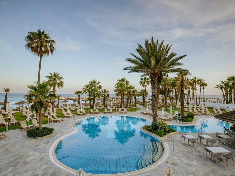 Poolanlage mit Palmen, weißen Sonnenschirmen und Liegestühlen, blauer Himmel im Hintergrund.