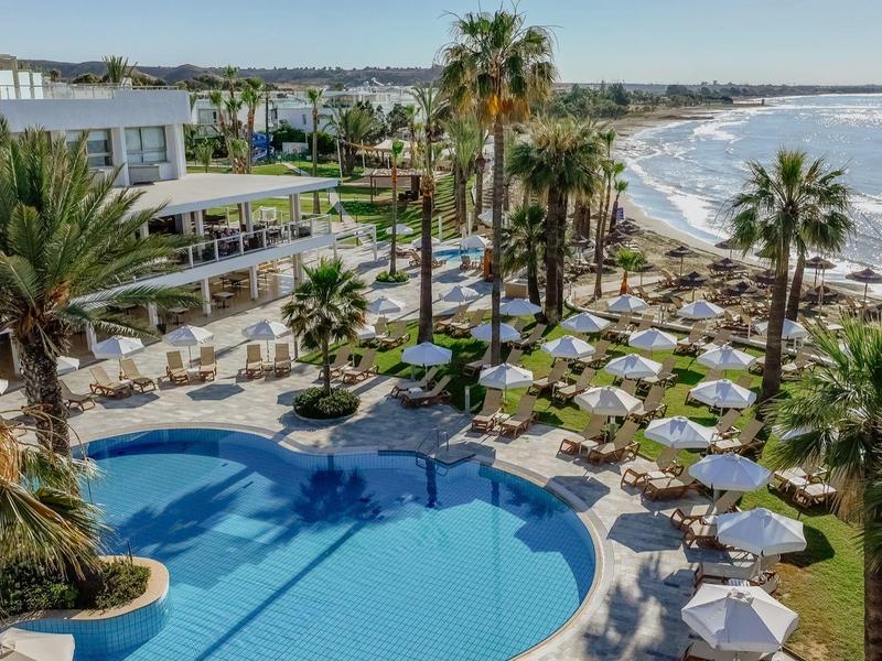Großer Hotelpool mit blauer Wasserfläche, umgeben von Liegen, Palmen und Blick auf das Meer.