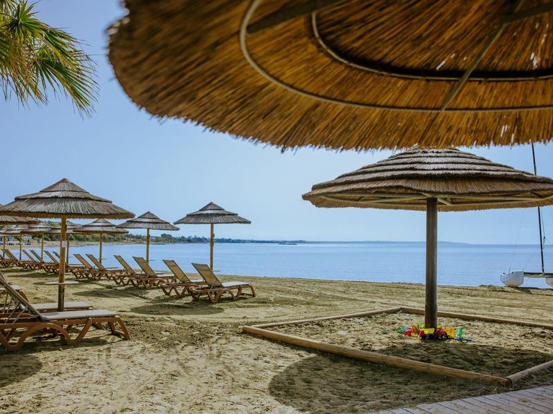 Strand mit Sand, Holzliegen und mehreren Strohhut-Sonnenschirmen am ruhigen blauen Meer.
