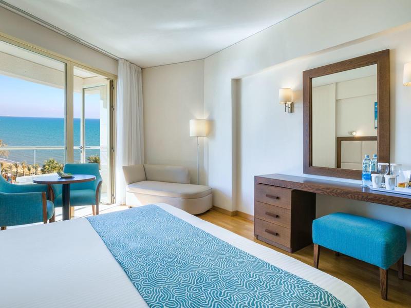 Helles Hotelzimmer mit Meerblick, blauem Bettläufer, zwei Sesseln und kleinem Schreibtisch.