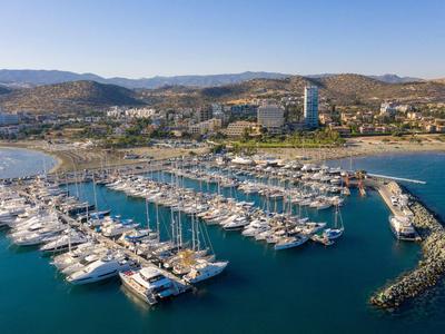 A marina filled with boats and yachts set against a coastal town and hills under clear sky.