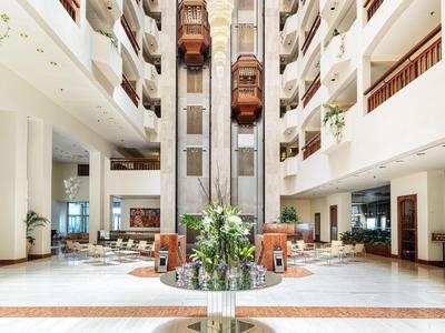 Bright hotel lobby with glass elevators and plants in a spacious seating area.