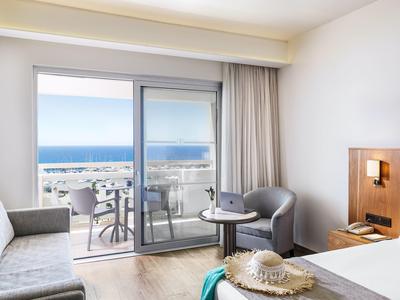 Hotel room with ocean view, balcony, sofa, chair, and small table with hat