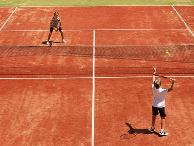Dos jugadores juegan tenis en una pista de arcilla roja bajo luz solar brillante.