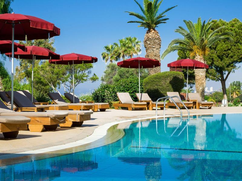 Poolside sun loungers under red umbrellas surrounded by palm trees on a sunny day.