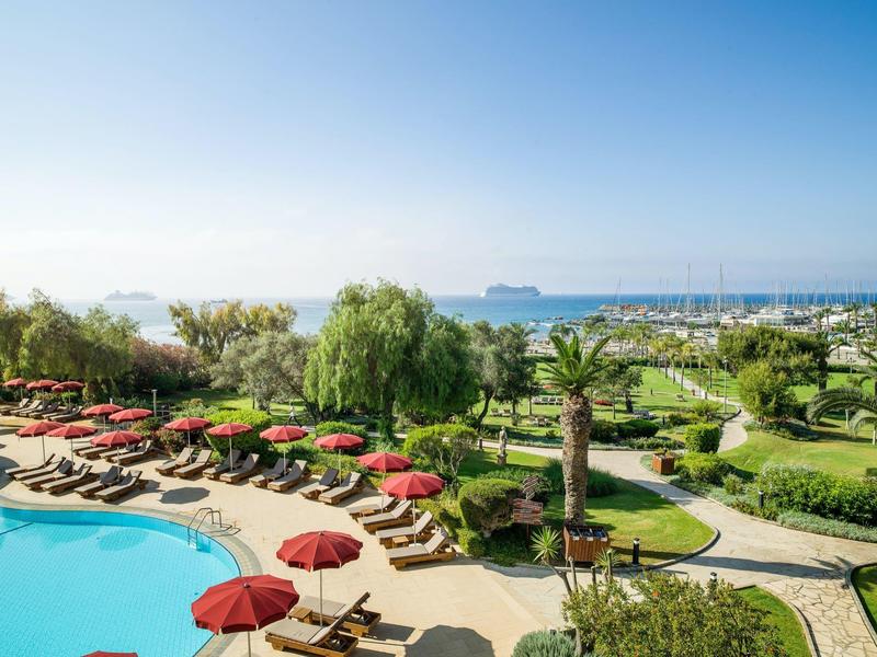 Resort pool area with sun loungers and red umbrellas near greenery and ocean.