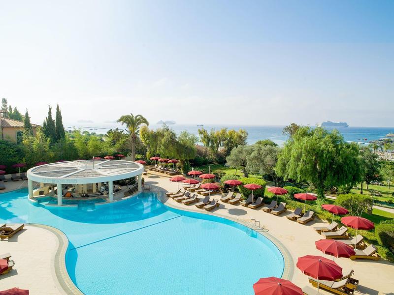 Curved outdoor pool with red umbrellas and sunbeds near green trees and sea view.