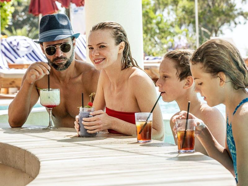 Four people relaxing by a poolside holding drinks on a sunny day.