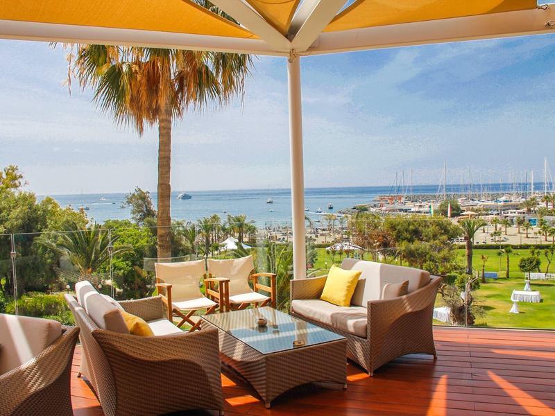 Cozy outdoor seating on a terrace overlooking palm trees and a marina by the sea.