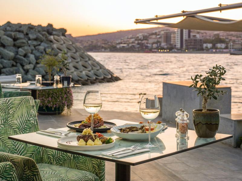 A table set with food and wine by the water during sunset with a rocky shore and buildings in the background.