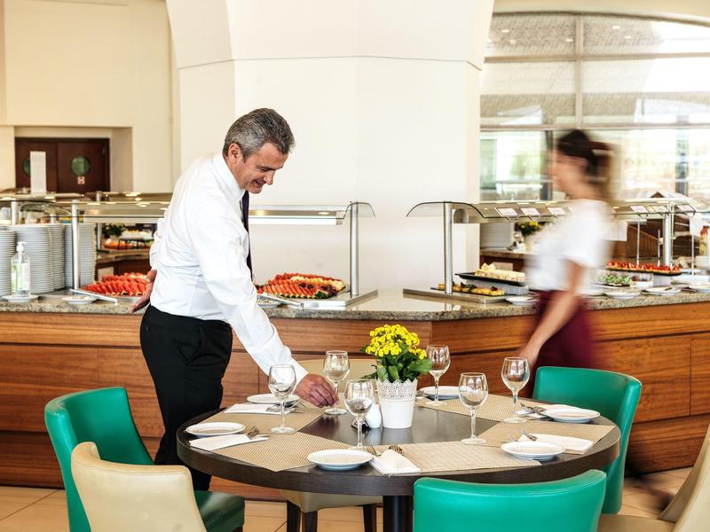 Hotel dining area with round table, teal chairs, waiter serving, buffet background.