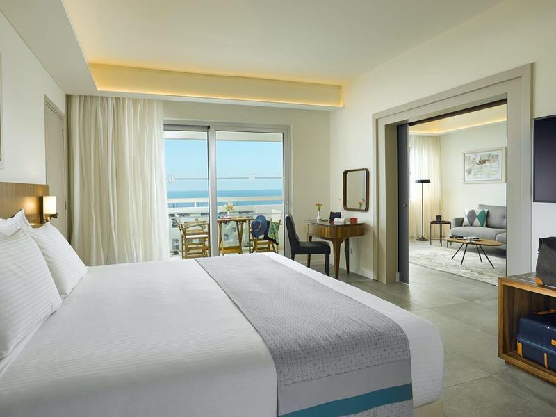 Modern hotel room with large bed, balcony view of city and sea, and adjoining living area.