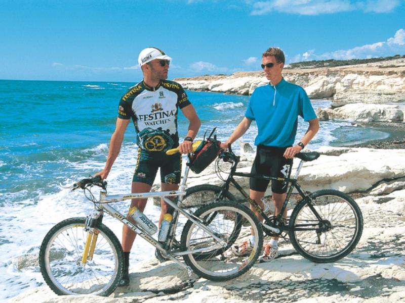 Zwei Radfahrer mit Fahrrädern am felsigen Meerufer bei sonnigem Wetter.