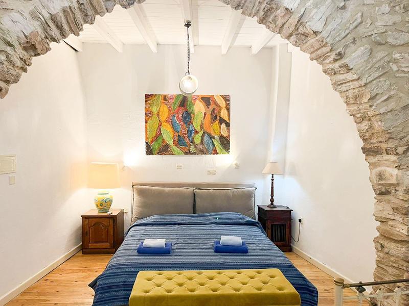 Schlafzimmer mit Doppelbett unter einem Steinbogen, Garderoben mit Nachttischen und Lampen, bunte Wandkunst.